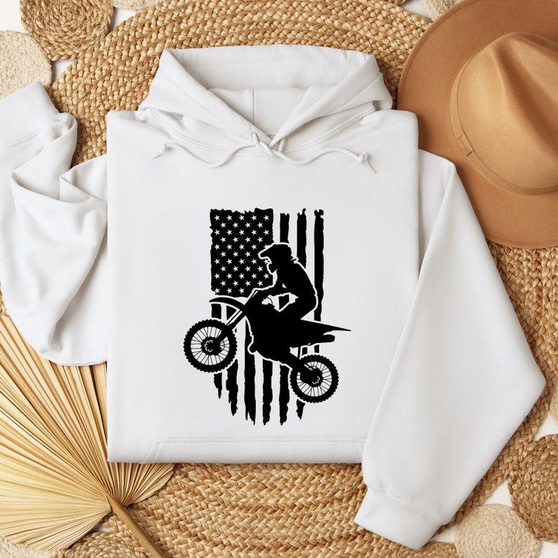 May include: White hooded sweatshirt with a black silhouette of a motocross rider jumping over a distressed American flag.