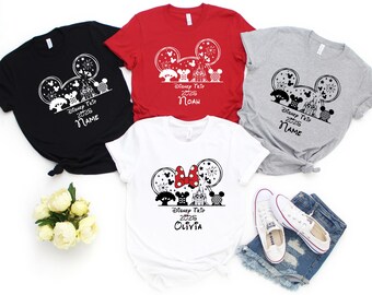 May include: Four t-shirts in black, red, gray, and white, each featuring a Disney-themed graphic. The design includes Mickey Mouse ears, a castle, and the text "Disney Trip 2025" or "2026" with a name. A pair of white sneakers and denim shorts are also visible.