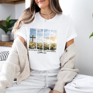 May include: White t-shirt with a graphic of a wooden cross in a field of flowers. The text "He is not here. He has risen. Matthew 28:6" is printed below the graphic.