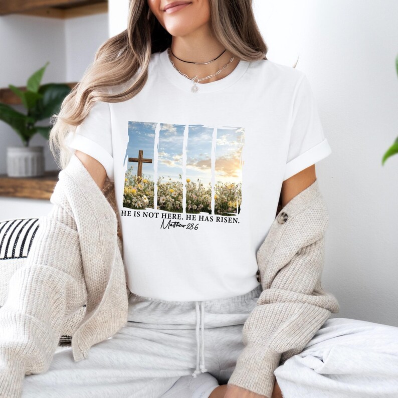 May include: White t-shirt with a graphic of a wooden cross in a field of flowers. The text "He is not here. He has risen. Matthew 28:6" is printed below the graphic.