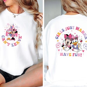 Puede incluir: Sudadera blanca con un gr&aacute;fico rosa y morado de Minnie Mouse y Daisy Duck. La parte delantera de la camiseta dice "Can't Leave" y la parte trasera dice "Girls Just Wanna Have Fun!"