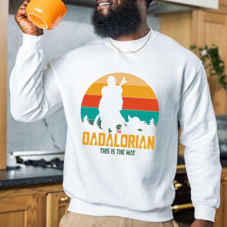 Puede incluir: Sudadera blanca de manga larga con un dise&ntilde;o gr&aacute;fico. El dise&ntilde;o presenta el texto "The Dadalorian" y "This is the Way" con una silueta sobre un fondo de puesta de sol en tonos turquesa, naranja y amarillo.