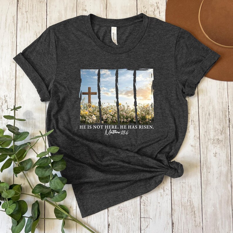 May include: A dark gray t-shirt with a white graphic of a cross in a field of flowers. The graphic is surrounded by a white frame with black lines. The text "He is not here. He has risen. Matthew 28:6" is printed below the graphic.