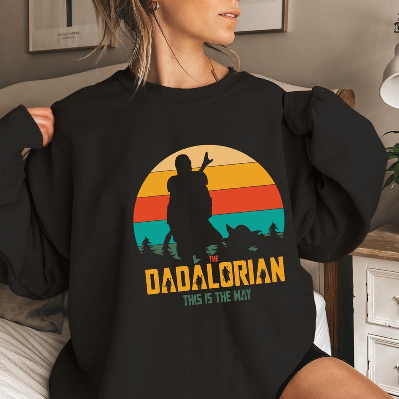 Puede incluir: Sudadera negra con un dise&ntilde;o gr&aacute;fico de una silueta contra un atardecer. El texto "The Dadalorian" y "This is the Way" es amarillo, con un estilo retro.