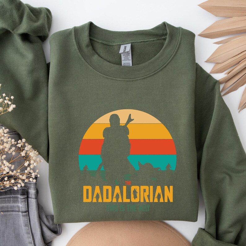 Puede incluir: Sudadera verde oliva con un gr&aacute;fico retro de puesta de sol. El dise&ntilde;o presenta la silueta de una figura con la mano levantada, con el texto "THE DADALORIAN" y "THIS IS THE WAY" en amarillo. Un estilo casual.