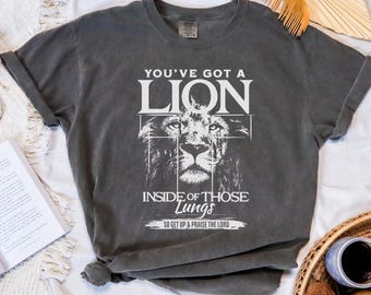 May include: Dark gray t-shirt with the text "YOU'VE GOT A LION INSIDE OF THOSE LUNGS" and a lion graphic. The shirt has rolled-up sleeves and a crew neck. The text "SO GET UP & PRAISE THE LORD" is also printed on the shirt.