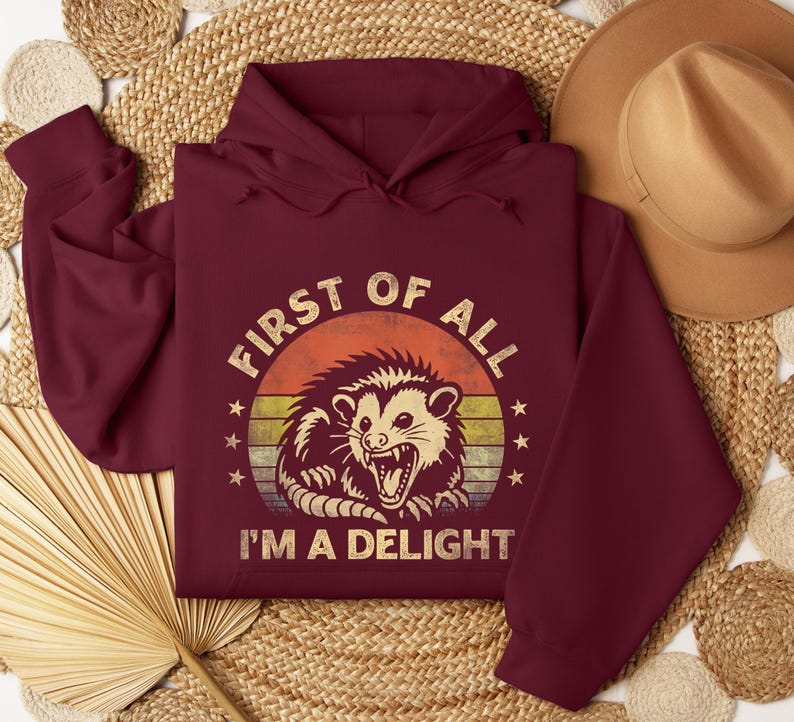 May include: Burgundy hoodie with a distressed graphic of an opossum inside a circle with the text "FIRST OF ALL I'M A DELIGHT". The design features a retro sunset color scheme. The hoodie is displayed on a woven surface.