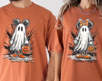 May include: Two orange t-shirts with Halloween-themed designs. Each shirt features a cartoon ghost with mouse ears, holding a jack-o'-lantern bucket. The ghost is white, with black accents and orange shoes. The background has black splatter effects.