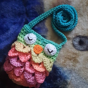 Puede incluir: Un peque&ntilde;o bolso de ganchillo en forma de b&uacute;ho con un cuerpo de colores arco&iacute;ris y una correa turquesa. El b&uacute;ho tiene ojos blancos con pupilas negras y un pico naranja.