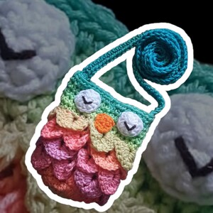 Puede incluir: Una peque&ntilde;a bolsa de ganchillo con forma de b&uacute;ho con un cuerpo de colores arco&iacute;ris y una correa turquesa. La bolsa tiene ojos blancos con pupilas negras y un pico naranja brillante.