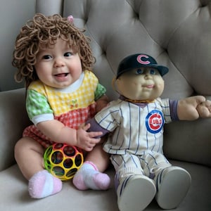 May include: A baby wearing a brown yarn wig and a yellow, green, and red checkered outfit, holding a colorful multi-holed plastic ball, sitting next to a Cabbage Patch Kids doll dressed in a white and blue pinstriped Chicago Cubs baseball uniform.