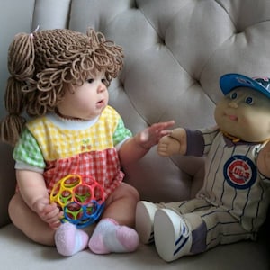 May include: A baby wearing a brown yarn wig and a yellow, green, and red checkered outfit, holding a colorful multi-holed plastic ball. Beside the baby sits a Cabbage Patch Kids doll wearing a Chicago Cubs baseball uniform and a blue cap.