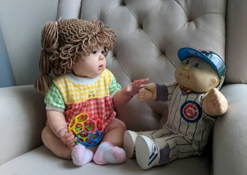 Peut inclure: Un b&eacute;b&eacute; portant une perruque en laine brune et une tenue &agrave; carreaux jaunes, verts et rouges, tenant une balle en plastique color&eacute;e &agrave; trous. &Agrave; c&ocirc;t&eacute; du b&eacute;b&eacute; se trouve une poup&eacute;e Cabbage Patch Kids v&ecirc;tue d'un uniforme de baseball des Chicago Cubs.