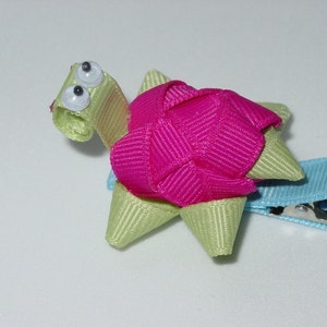 May include: A green and pink ribbon turtle hair clip with googly eyes. The clip is attached to a light blue ribbon.