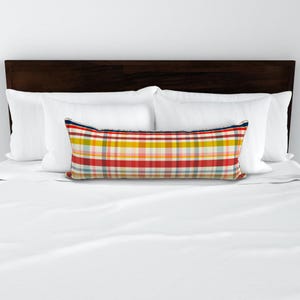May include: A rectangular lumbar pillow with a colorful plaid pattern in red, yellow, blue, and white. The pillow is placed on a white bed with white pillows and a dark brown headboard. The pillow is approximately 30 inches long.