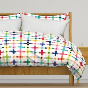 May include: A bed with a wooden frame and white pillows. The bedding features a colorful plaid pattern with intersecting lines and dots in red, blue, green, yellow, and purple on a white background. The overall theme is a bright and cheerful bedroom.