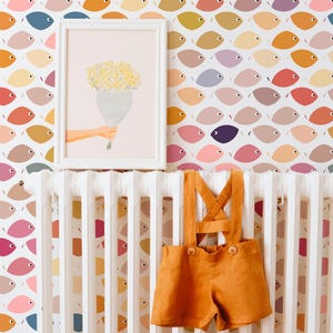 May include: An orange linen romper with wooden buttons hangs on a white crib. A framed print of a hand holding a bouquet of yellow flowers is above the crib. The wallpaper features a repeating pattern of colorful fish.