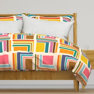 May include: A white duvet cover with a geometric pattern of colorful squares and stripes. The duvet cover is on a bed with a wooden headboard and two white pillows.