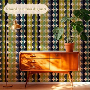 May include: A mid-century modern wooden cabinet with three drawers and a single door, topped with a white surface. A potted plant sits on top of the cabinet. The background features a wallpaper with a repeating pattern of green, blue, and cream circles. A gold floor lamp stands to the left. Text reads: "beloved by interior designers."