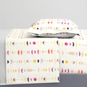 May include: White bed sheets and a pillow with a colorful, repeating pattern of abstract shapes in pink, yellow, purple, and orange. The sheets are neatly folded, and the pillow is placed on top.