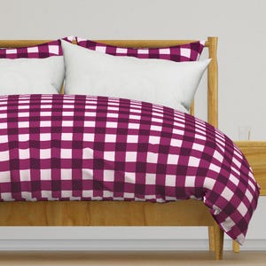 May include: A bed with a wooden frame and a duvet cover featuring a large, checkered pattern in shades of purple and white. Two white pillows are visible, along with a wooden nightstand.