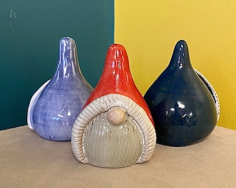 May include: Three ceramic figurines shaped like teardrops or gnomes. The center figure has a bright red pointed hat, a textured beige beard, and a small round nose. The left figure is light purple, and the right figure is a deep, glossy navy blue.