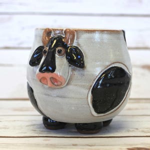 May include: A ceramic mug shaped like a cow with black and white spots. The mug has a brown nose, black eyes, and brown horns. The mug is sitting on a white wooden surface.