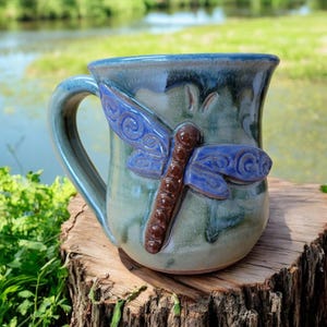 May include: A handmade ceramic mug with a blue and green glaze. A 3D dragonfly with blue wings and a brown body is attached to the mug. The mug is sitting on a wooden surface.