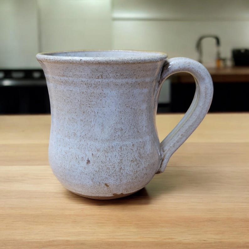 May include: A white ceramic mug with a rounded body and a handle. The mug has a speckled glaze and a slightly textured surface.