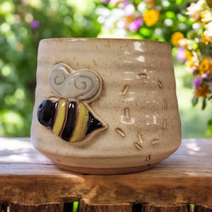 May include: A ceramic mug with a bee design. The mug is white with a brown glaze and has a bee with yellow and black stripes on the front. The bee is flying towards the right side of the mug.