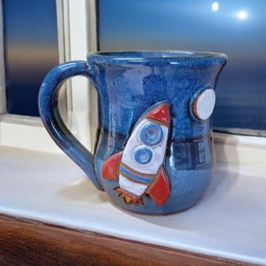 May include: A blue ceramic mug with a 3D rocket ship design. The rocket is red, white, and blue with a white moon on the side of the mug.