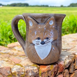 May include: A brown ceramic mug with a fox face sculpted on the front. The fox has a white bib and brown fur.