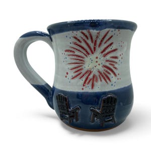 May include: Handmade ceramic mug with a blue, white, and red color scheme. The mug features a firework design in red and white, with two Adirondack chairs embossed on the base. The handle is white and blue.