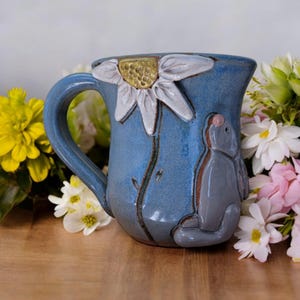 May include: A blue ceramic mug with a white daisy and a gray rabbit sculpted on the side. The mug has a handle and is sitting on a wooden surface.