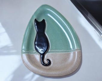 May include: A ceramic cat-shaped dish with a black cat silhouette. The dish is teardrop-shaped, with a light green upper section and a tan lower section. The cat is facing away, with its tail curled. A decorative item.