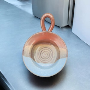 May include: A ceramic dish with a handle, featuring a blue, brown, and orange glaze. The dish has a circular shape and a slightly raised rim.
