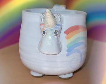 May include: White ceramic mug with a unicorn face and a rainbow painted on the side. The unicorn has a white horn and blue eyes.