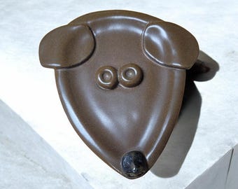 May include: A brown ceramic dog figurine with large ears and two black eyes. The dog's nose is black and the figurine is sitting on a white surface.