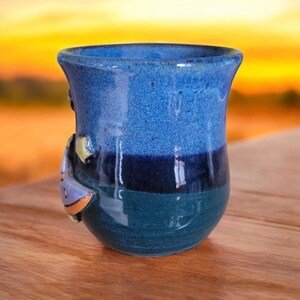 K&ouml;nnte beinhalten: Eine blaue und gr&uuml;ne Keramiktasse mit einem 3D-Design eines Segelboots an der Seite. Die Tasse hat eine glasierte Oberfl&auml;che und ist handgefertigt.