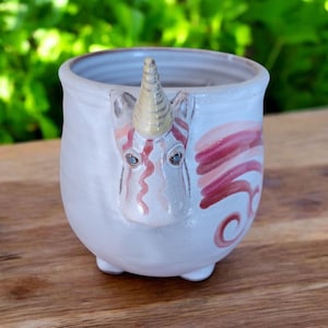 May include: White ceramic mug with a unicorn face and pink and white stripes. The unicorn has a yellow horn and blue eyes.