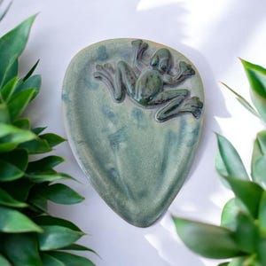 May include: A green ceramic dish with a frog design. The dish is shaped like a leaf and has a glazed finish.
