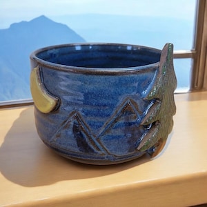 May include: A blue ceramic bowl with a mountain and moon design. The bowl has a green tree-shaped handle.