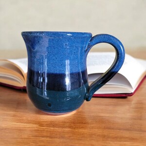 K&ouml;nnte beinhalten: Eine blaue und gr&uuml;ne Keramiktasse mit abgerundetem K&ouml;rper und Henkel. Die Tasse hat eine glasierte Oberfl&auml;che und ist mit Streifen in Blau, Dunkelblau und Gr&uuml;n verziert.