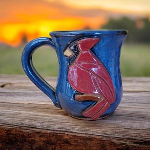 May include: A blue ceramic mug with a red cardinal bird design. The mug has a handle and is sitting on a wooden surface.