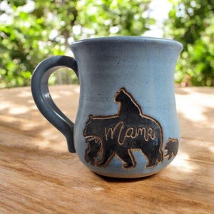 May include: A blue ceramic mug with a black bear family design. The mug features a bear silhouette with the word "Mama" and two smaller bear cubs. The handle is a matching blue color.