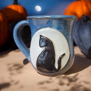 May include: A blue ceramic mug with a white moon and a black cat silhouette. The mug has a rounded handle.
