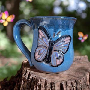 May include: A blue ceramic mug with a butterfly design. The butterfly is in shades of purple, pink, and black. The mug has a handle and is sitting on a tree stump.
