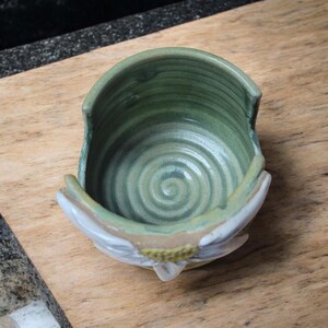 May include: A green ceramic bowl with a white and yellow floral design. The bowl has a swirl pattern on the inside and a small opening on the side.