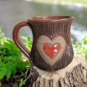 May include: A brown ceramic mug with a tree bark texture and a red heart design. The mug is sitting on a tree stump.