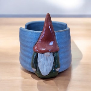 May include: A blue ceramic planter with a gnome design. The gnome has a red hat, a white beard, and a green shirt.